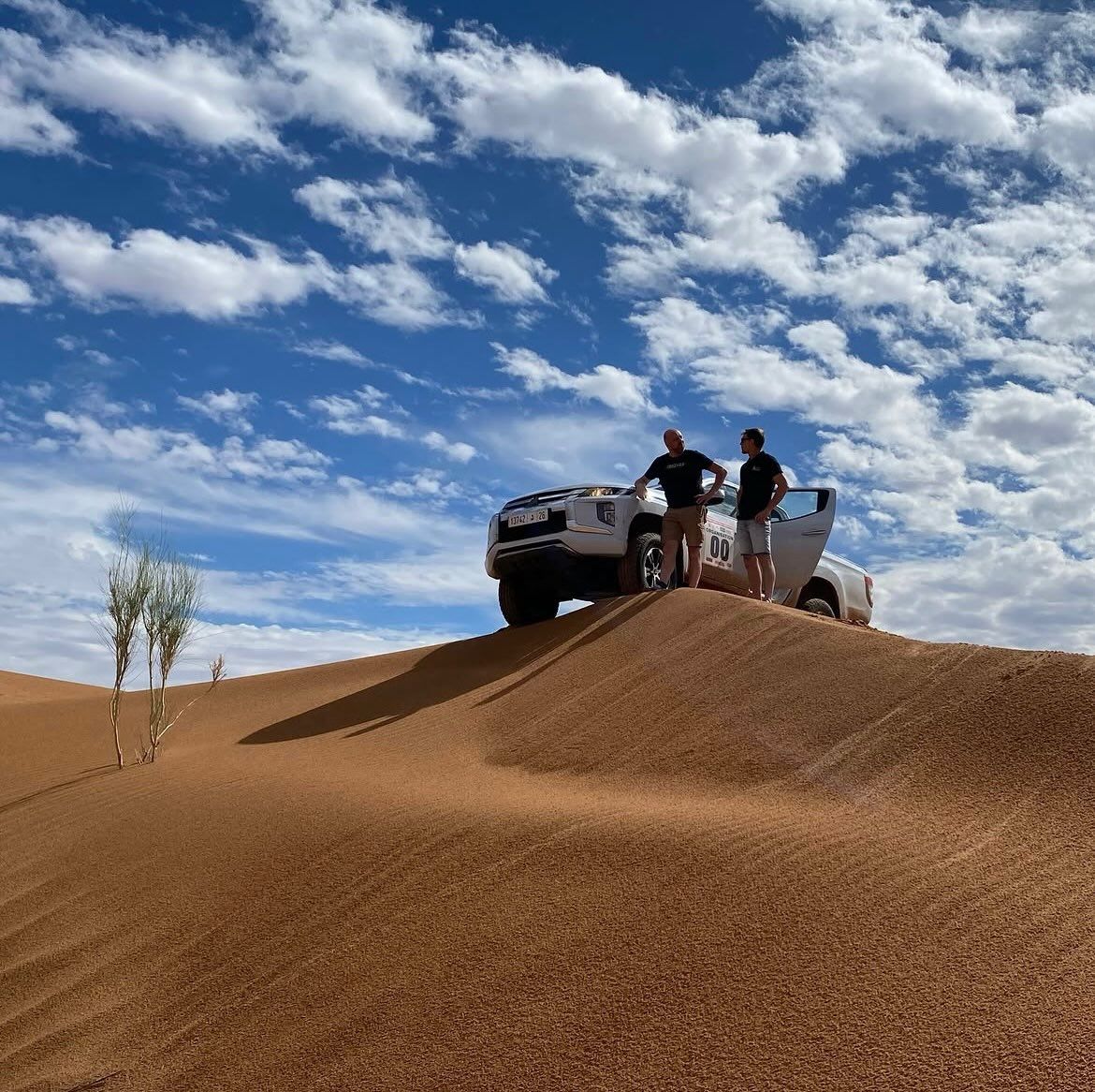 Peter met de witte volgwagen bovenop een zandduin, onder een blauwe lucht met wolken