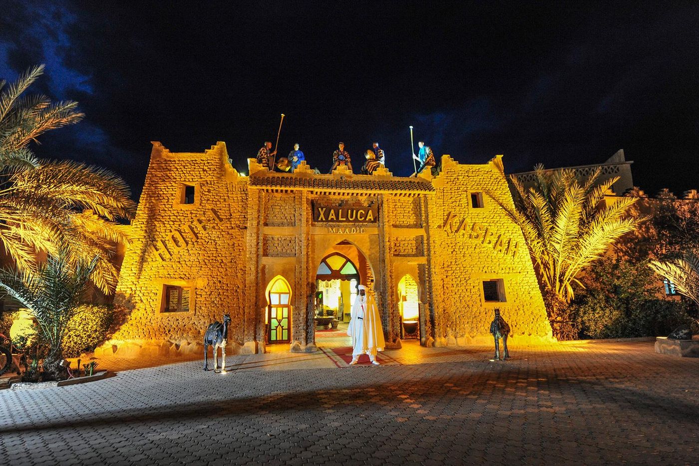 Verlichte toegangspoort van Hotel Kasbah Xaluca Maadid bij nacht, tussen palmbomen