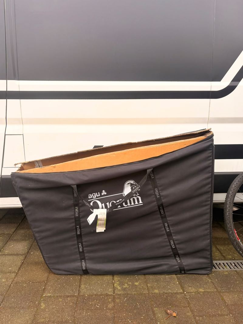 Zwarte fietstas met een kartonnen fietsdoos erin, tegen een witte bestelwagen